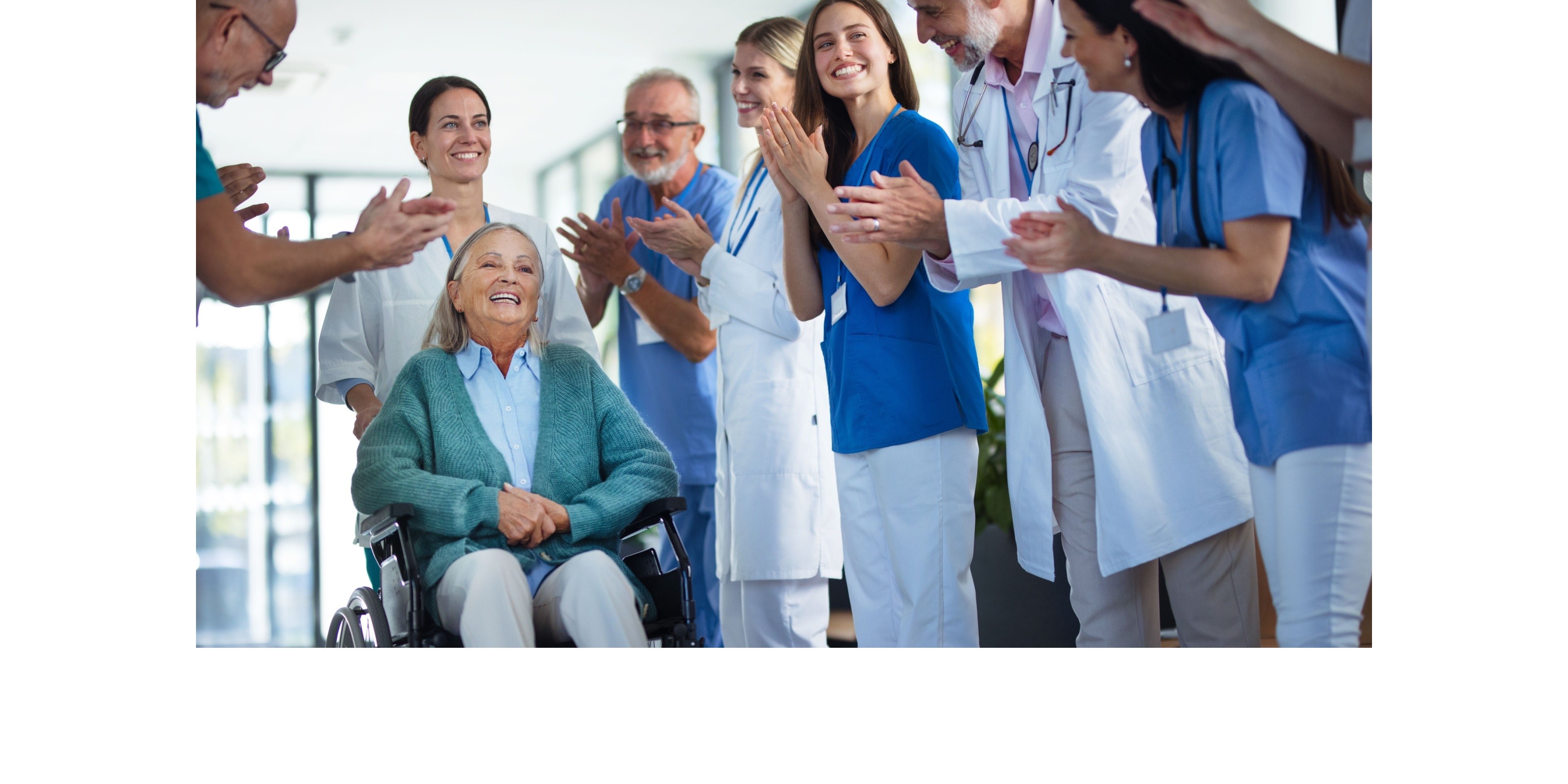 Smiling elderly woman in wheelchair surrounded by applauding medical staff in a hospital hallway