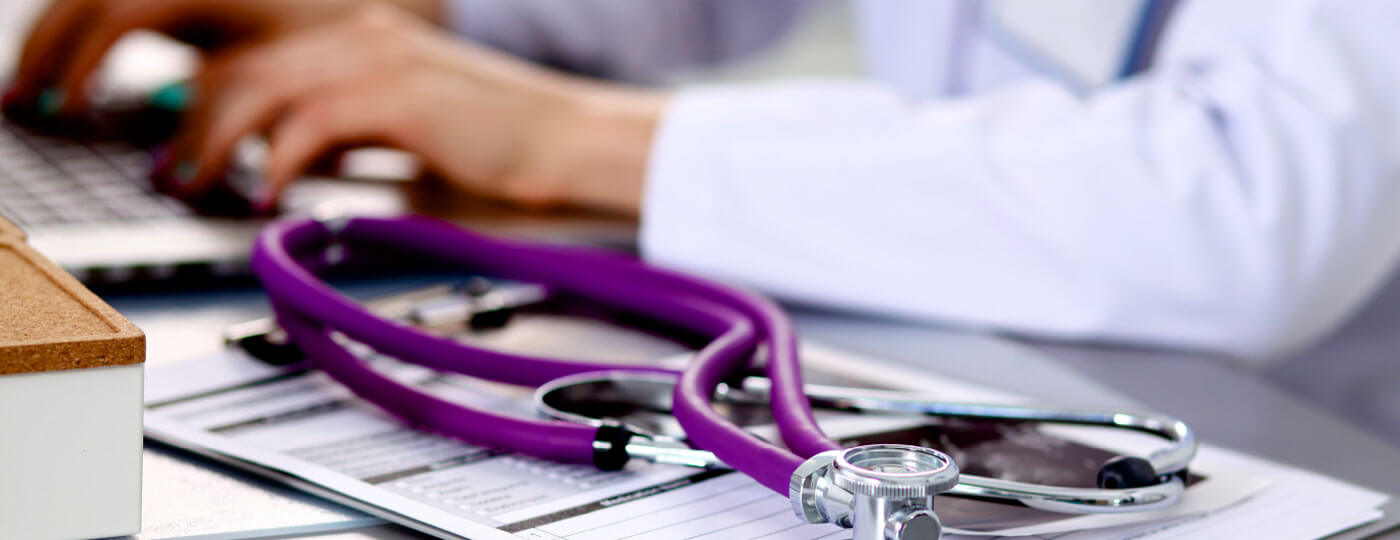 Close-up of a purple stethoscope on a medical chart with a healthcare professional typing on a laptop in the background