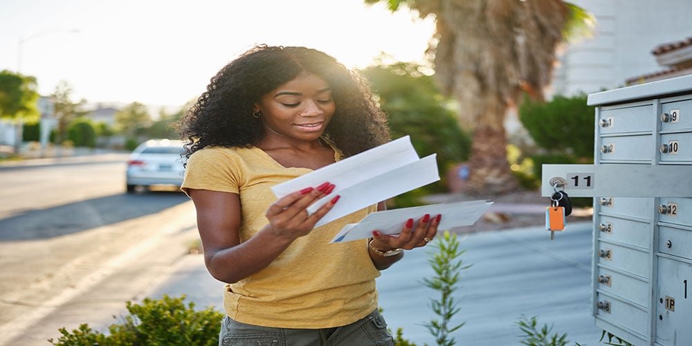 Smiling woman checking her mail at an outdoor community mailbox on a sunny day.