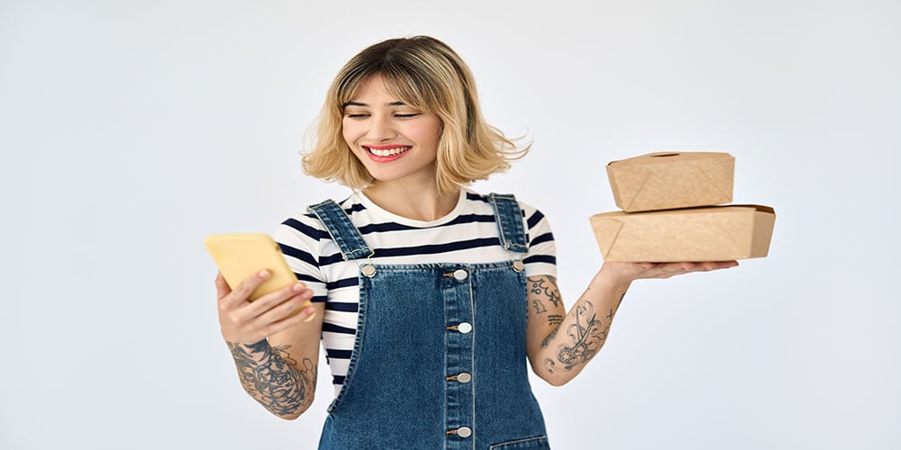 Happy woman holding two takeout food containers and looking at her smartphone.